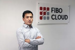 Өөртөө итгэлтэй бизнесмэн "FIBO CLOUD" гэсэн хананд наасан тэмдгийн өмнө гараа зөрүүлэн зогсож байна. Тэрбээр цайвар цэнхэр өнгийн товчтой цамц өмссөн байна. Энэхүү тэмдэг нь түүний компанийн хэтийн төлөвийг илэрхийлсэн улаан, саарал дөрвөлжин бүхий дижитал сэдэвт лого агуулсан. - Business.MN