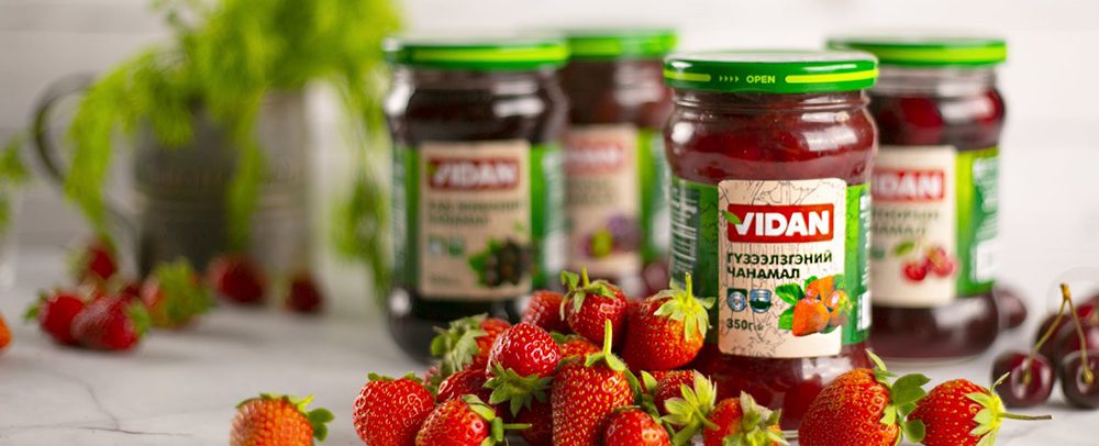 Jars of Vidan strawberry preserve are placed on a marble surface, surrounded by fresh strawberries. The jars have green caps and colorful labels. A leafy plant is blurred in the background.