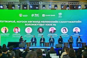 A panel discussion at a conference, sponsored by Тос Хаан Банк, features seven speakers on stage with microphones. Behind them, a large screen displays their names and titles in a non-English language. The engaged audience is visible in the foreground, eagerly absorbing the dialogue.