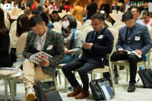 People are sitting on chairs in a conference room, reading materials. Some are wearing name tags and others have bags by their feet. The room has a neutral color scheme, and there are more individuals gathered in the background.