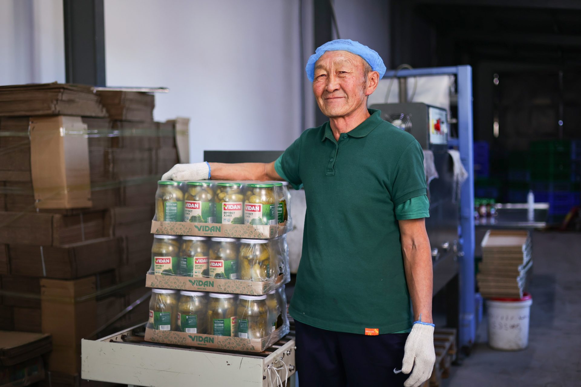 A smiling person wearing a blue hairnet, green shirt, and white gloves stands beside a stack of packaged goods in a warehouse. The packages are labeled "VIDAN" and likely contain bottled products. Boxes are seen in the background.