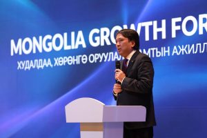 A man in a suit speaks at a podium against a blue background. The backdrop displays text reading "Mongolia Growth For" and additional text in another language.