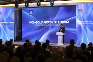 A speaker at a podium addresses an audience at the Mongolia Growth Forum. The backdrop displays the forum's theme: "Trade, Investment and Collaboration," with digital blue graphics and official logos.