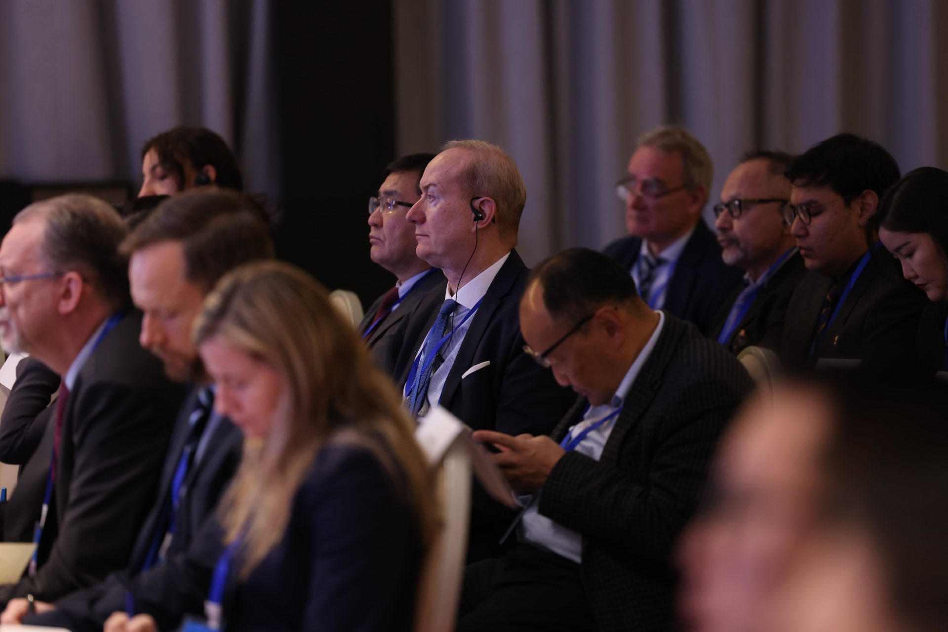 A group of people seated in a conference room, wearing formal attire. Some are listening attentively, and others are taking notes or using devices. Everyone is wearing name tags and conference lanyards.