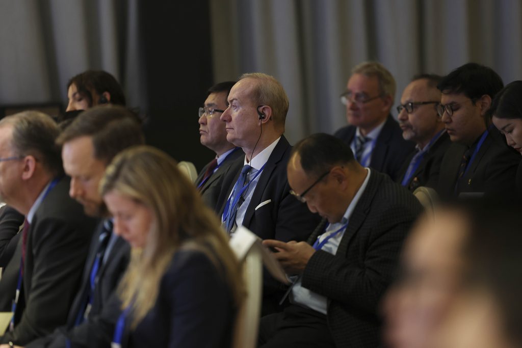 Audience members in formal attire are seated in rows, attentively listening to a presentation. Some wear headphones for translation, while others take notes. The room has neutral tones and a professional atmosphere.