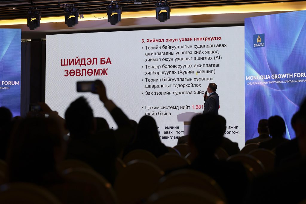 A speaker presents information on a screen at an event. The audience is seated, and the setting appears to be a conference room. The slide contains text in a non-English language, possibly relating to technology or governance.