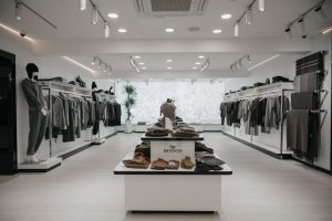 Modern clothing store interior with white walls and sleek lighting. Clothes and accessories are displayed neatly on racks along the walls and on tables in the center. A sign labeled "BOIDOS" is on a table with folded garments.