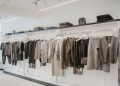 A clothing store interior showcasing a neatly arranged rack of neutral-toned sweaters and coats. Shelves above display folded garments and a few handbags. The space is well-lit with a minimalist, modern design.