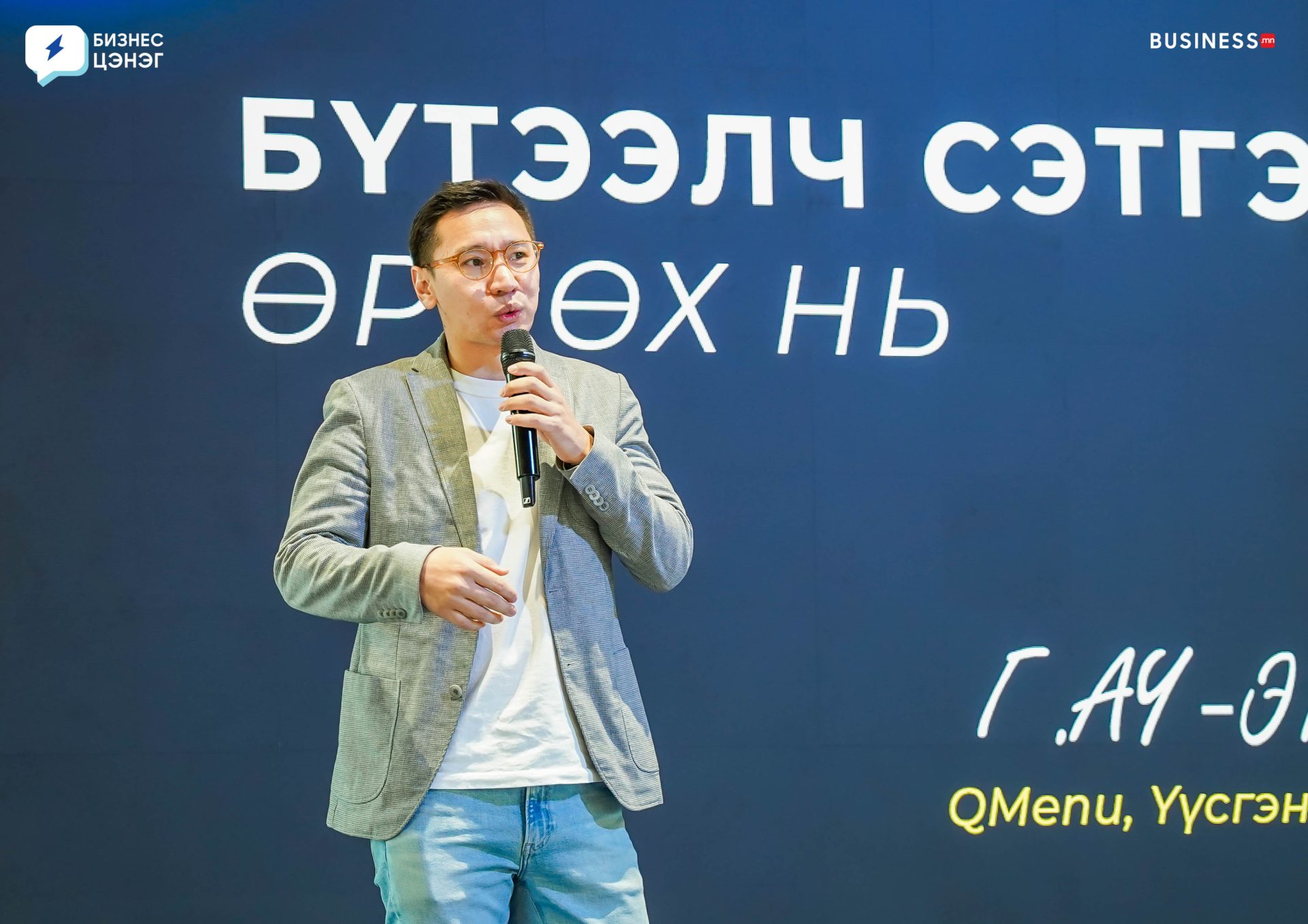 A person holding a microphone stands on stage in front of a dark screen with Mongolian text. They are wearing glasses, a gray blazer, and jeans. Logos are visible in the corners of the image.