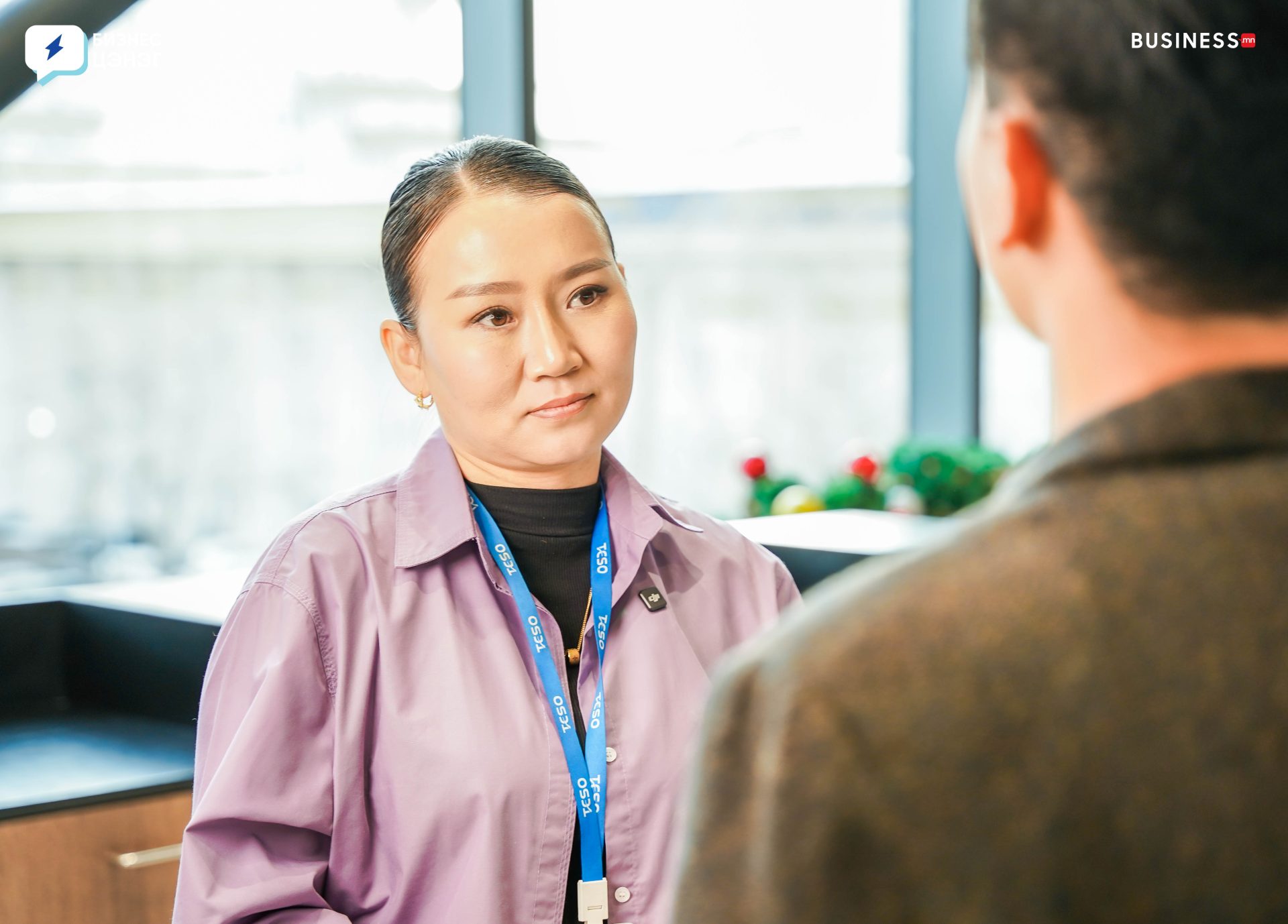 A person with a thoughtful expression and a lanyard stands facing another person in an office setting. The background is softly blurred, showing windows and some greenery.