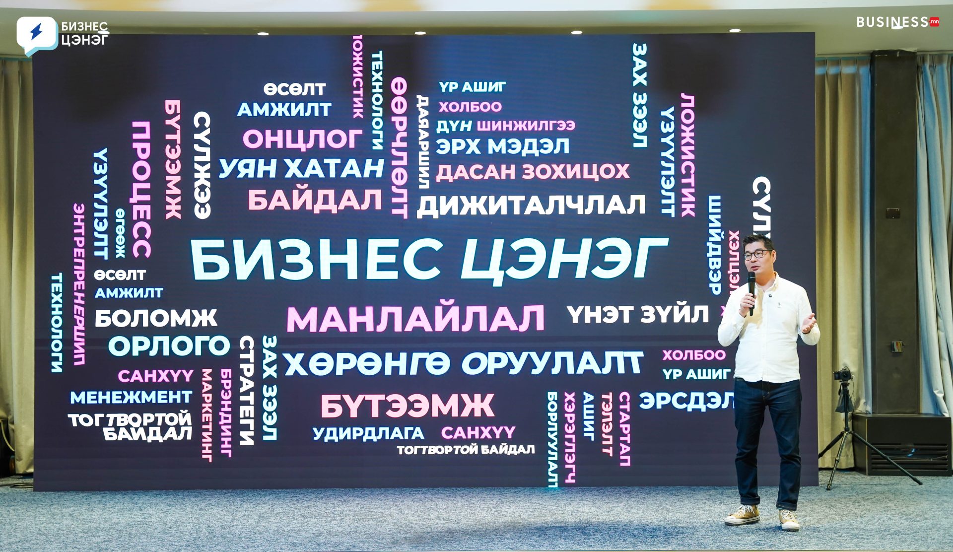 A speaker stands on stage next to a large screen displaying a colorful word cloud with Mongolian text. At this бизнес цэнэг уулзалт, the largest words include "БИЗНЕС ЦЭНЭГ," "МАНЛАЙЛАЛ," and "ХӨРӨНГӨ ОРУУЛАЛТ." The setting appears to be a dynamic business event.