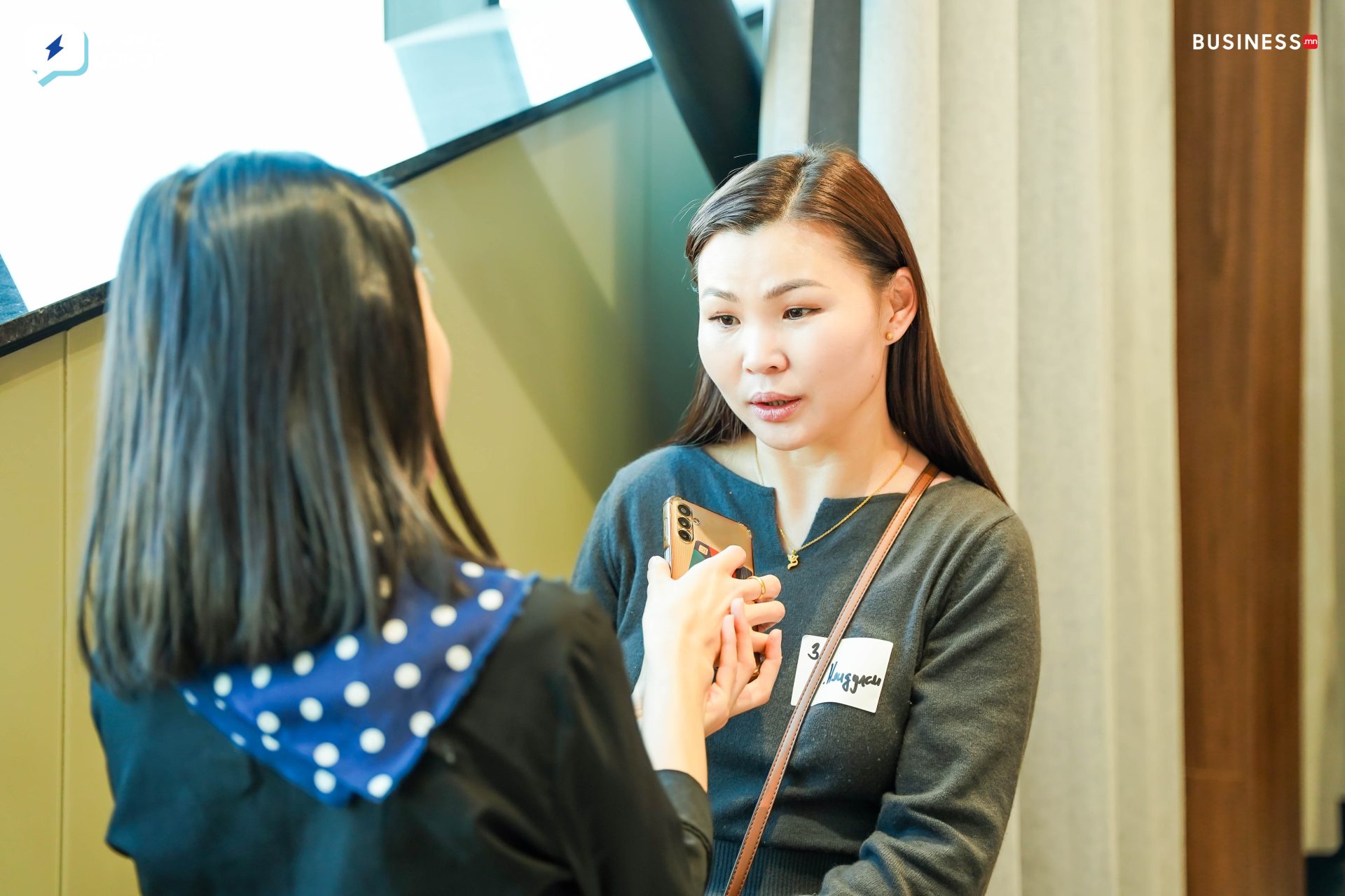 Two women are talking indoors. One is holding a smartphone as the other listens. Both are wearing name tags and casual clothing. A staircase is visible in the background.