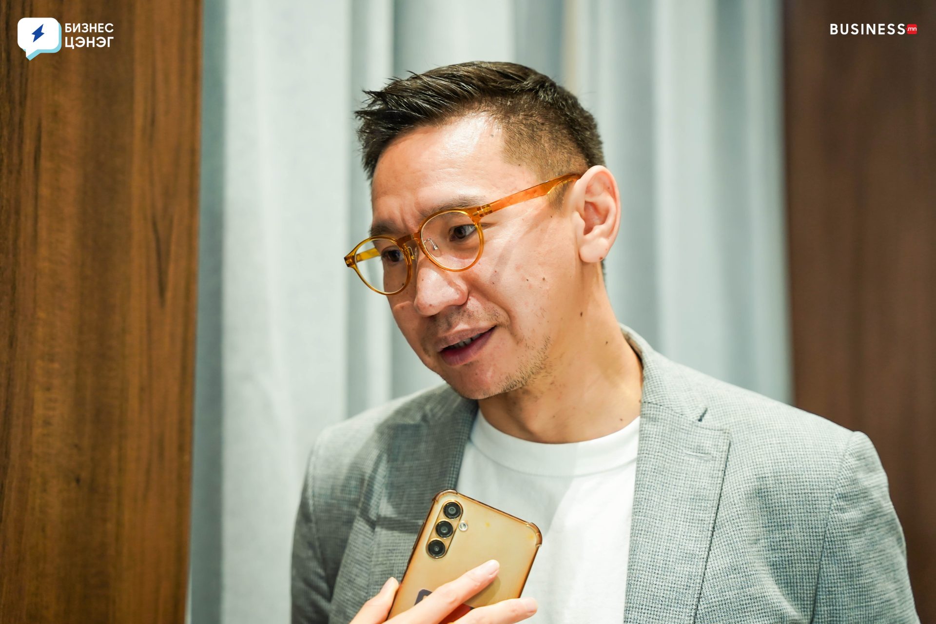 A man wearing glasses and a gray blazer is speaking, holding a smartphone. He stands in front of a wooden and curtain backdrop, with logos on the top corners of the image.