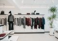 A modern clothing store features neatly arranged racks of grey and burgundy sweaters and jackets. A mannequin dressed in a grey sweater and black pants stands to the left. Bright lighting and a potted plant create a clean, stylish ambiance.