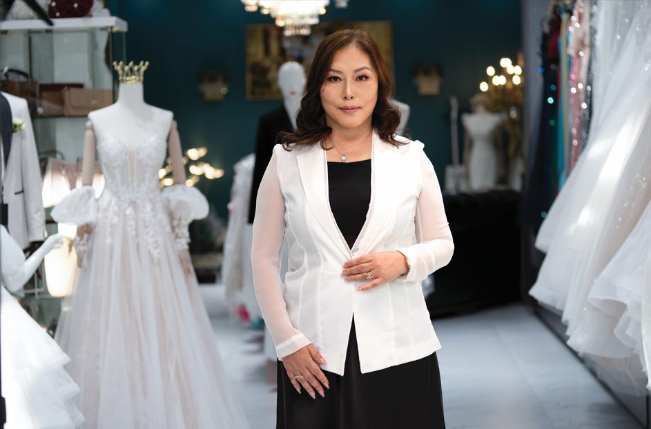 A woman stands confidently in a bridal shop, wearing a white blazer over a black dress. Behind her are elegant wedding gowns on mannequins, and the store is softly lit with chandeliers and decorative lighting.