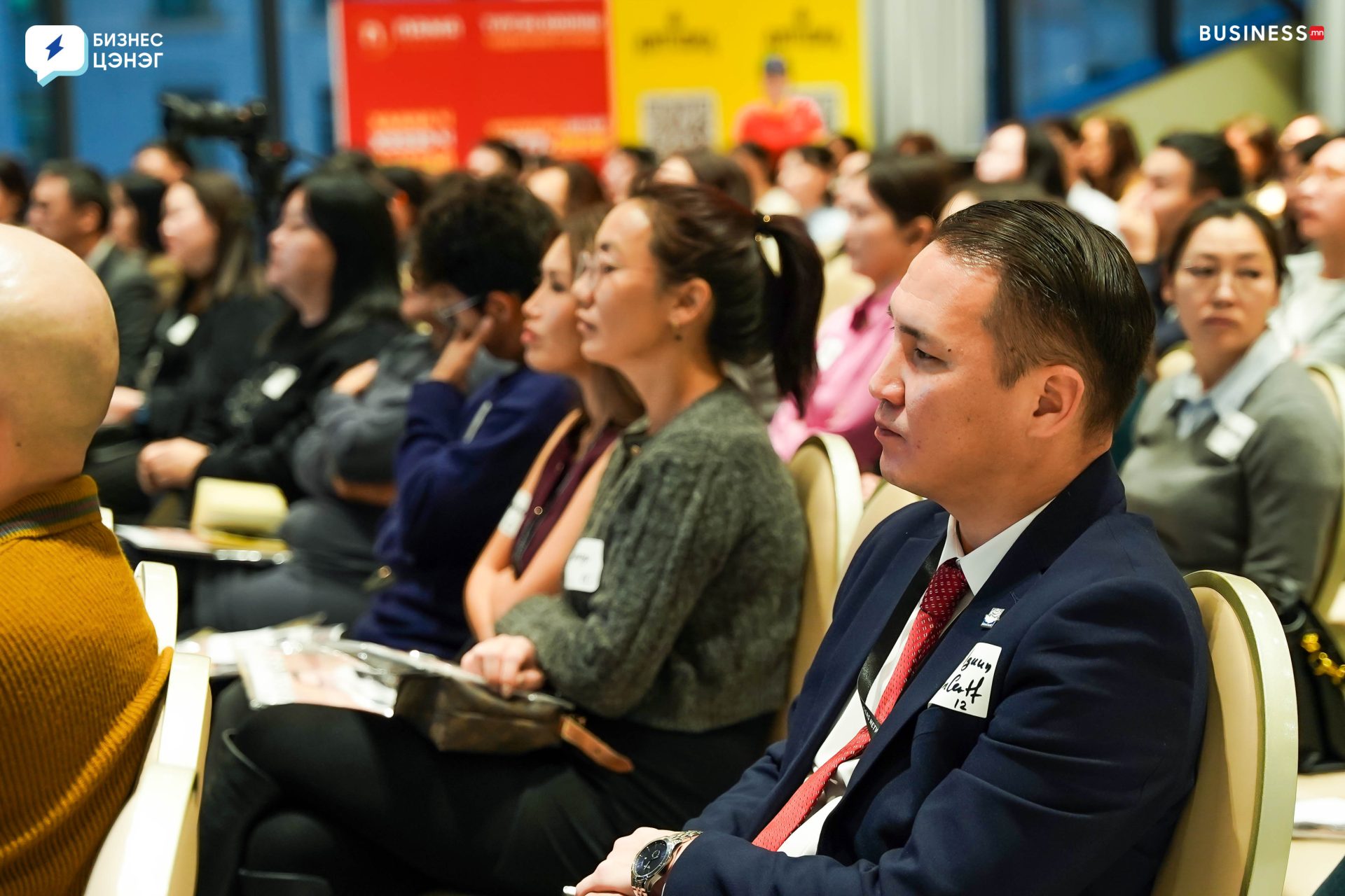 A diverse group of people seated in rows, attentively listening during a seminar or conference. A man in the foreground wears a suit and name tag. The room has colorful banners and bright lighting.