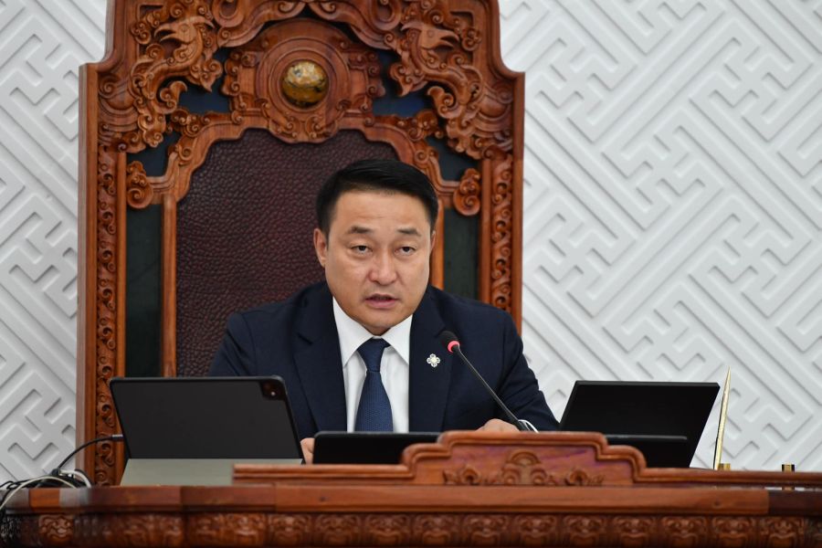 A man in a suit sits at an ornate wooden desk, speaking into a microphone. Behind him is a decorative carved wooden chair and a patterned white wall. Two laptops are open in front of him.