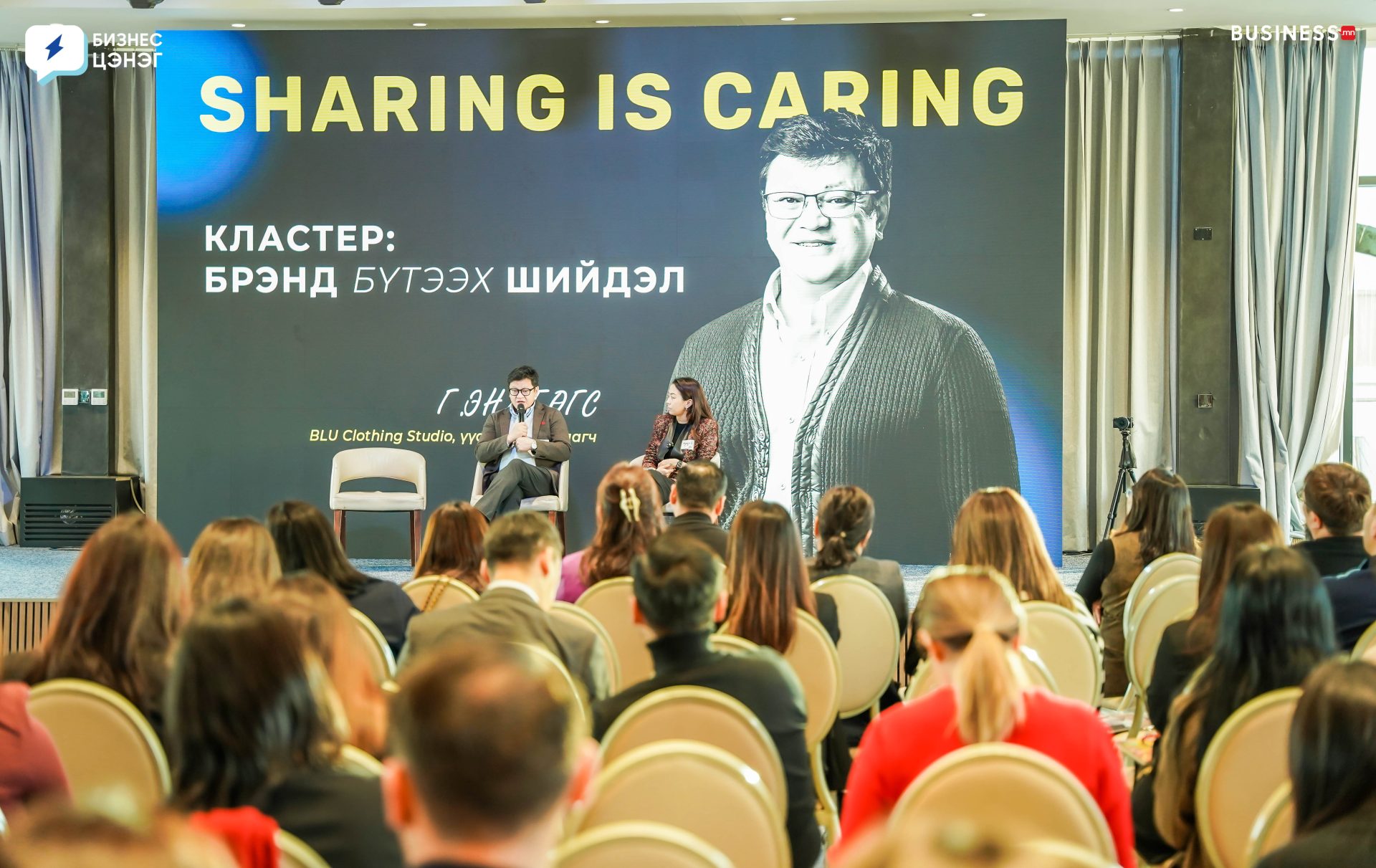 A large audience sits facing a stage with a presentation. The screen displays a speaker, a woman with short hair, and text reading "Sharing is Caring" and some additional information in another language. Two people sit on the stage.