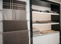 A display of folded, neutral-colored woven blankets and throws on shelves, with more blankets hanging on a wall rack. The patterns include plaid and solid designs, creating a cozy and inviting ambiance.