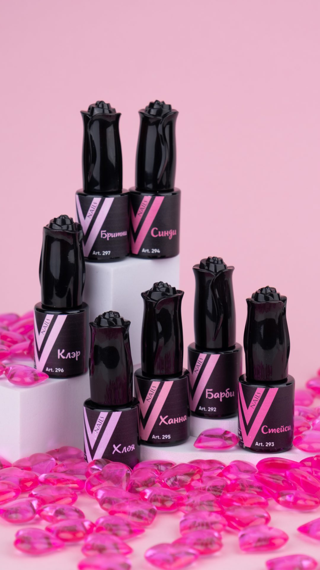 Eight black nail polish bottles with rose-shaped caps are arranged on white blocks against a pink background. The bottles have labels with names in a foreign script. Pink decorative stones are scattered around the display.