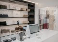 A modern retail store interior showcasing neatly stacked sweaters on shelves, alongside bags labeled "Bocados." A computer and payment terminal are situated on a white counter in the foreground. Soft fabrics in neutral tones hang on the right wall.