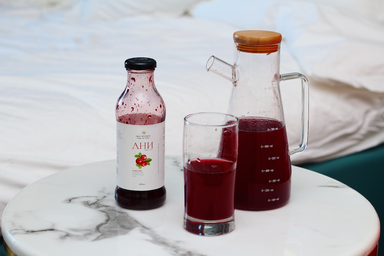 A bottle, a glass, and a carafe filled with red juice are placed on a white marble table. The carafe has a wooden lid, and a bed with white sheets is in the background.