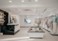 A minimalist clothing store with white walls and light wooden flooring. Neatly arranged neutral-toned clothes are displayed on racks and tables. A mannequin wearing a light green sweater stands by the entrance.