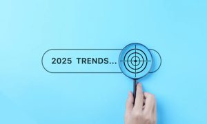 A hand holds a magnifying glass over the text "2025 TRENDS..." on a light blue background. The magnifying glass reveals a target icon over the word "TRENDS".