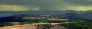 A panoramic view of rolling forested hills under a cloudy sky. Rain is visible in the distance, casting a soft shadow over the landscape. The foreground features a mix of green trees and open fields.