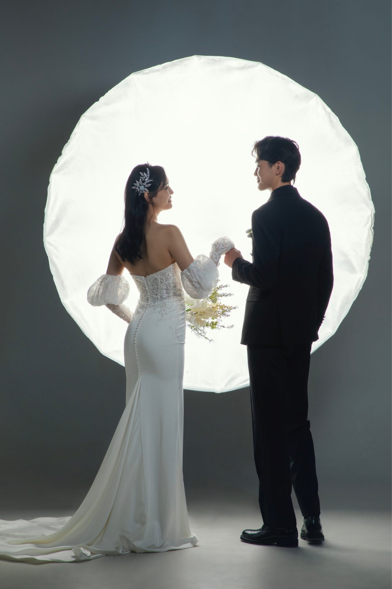 A bride in a white gown and a groom in a black suit stand holding hands in front of a bright circular light. The bride is holding a bouquet of flowers, and both are looking at each other, creating a romantic and elegant scene.