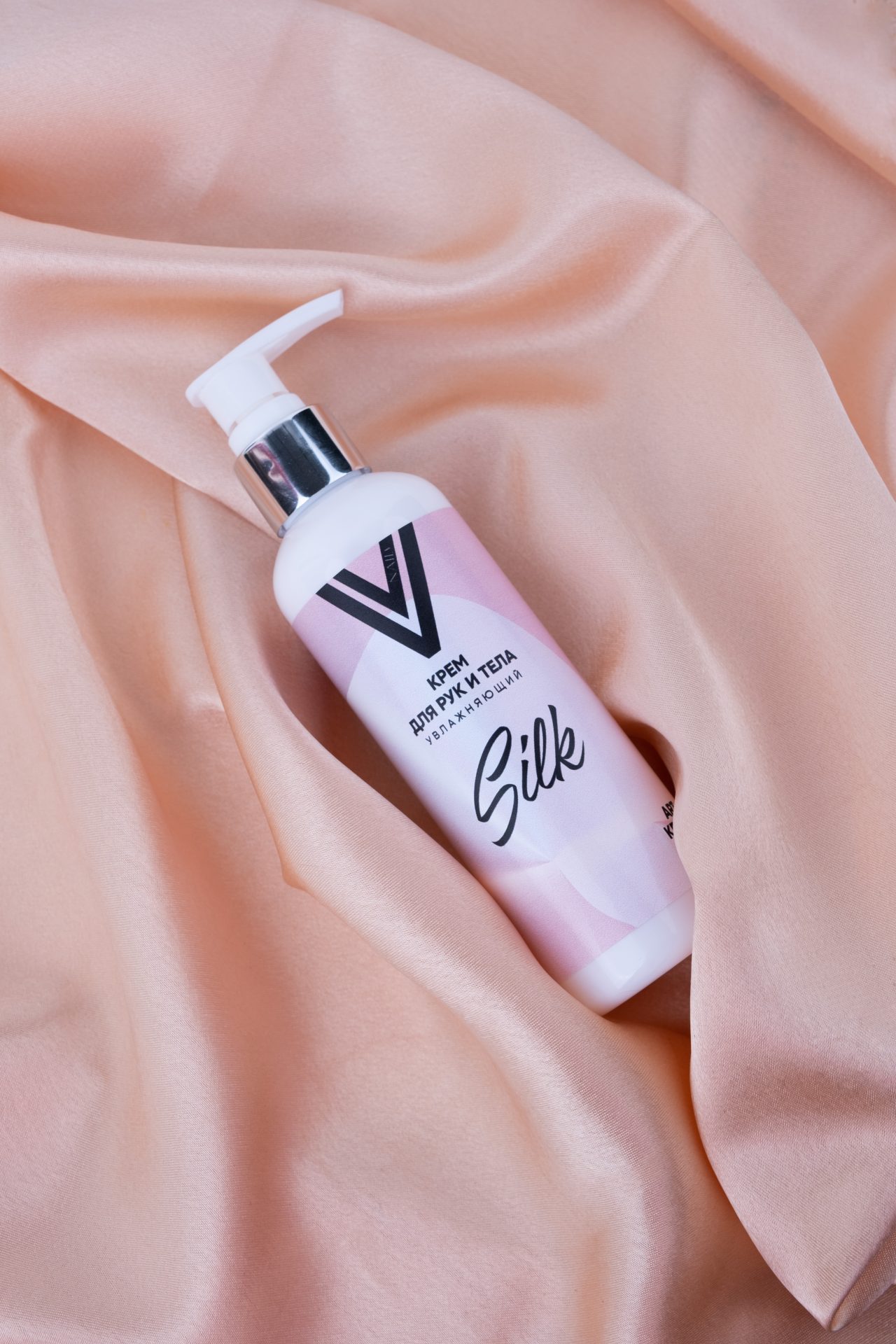 A bottle of body cream labeled "Silk" with a pump dispenser is placed on smooth, peach-colored satin fabric. The cream is packaged in a white and light pink container with a stylish design.