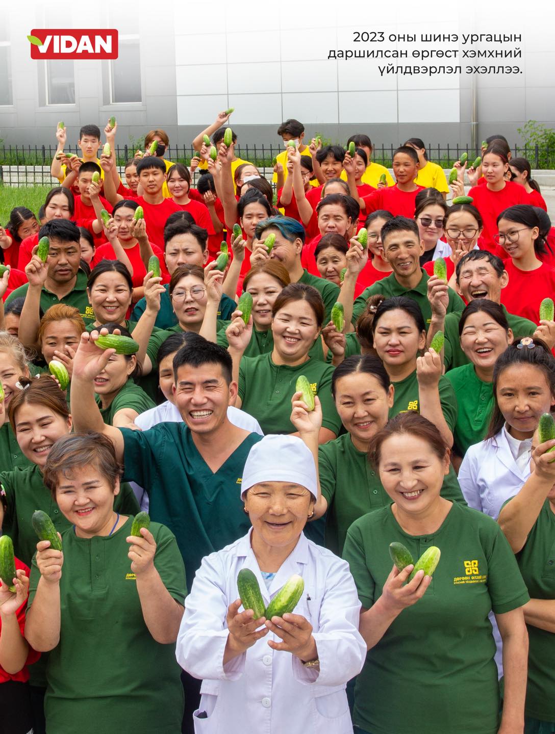 A large group of smiling people, some in colorful uniforms, hold up cucumbers in celebration. In the front center, a person in a white chef's outfit holds several cucumbers. The background features a modern building. Text in a non-English language is visible.