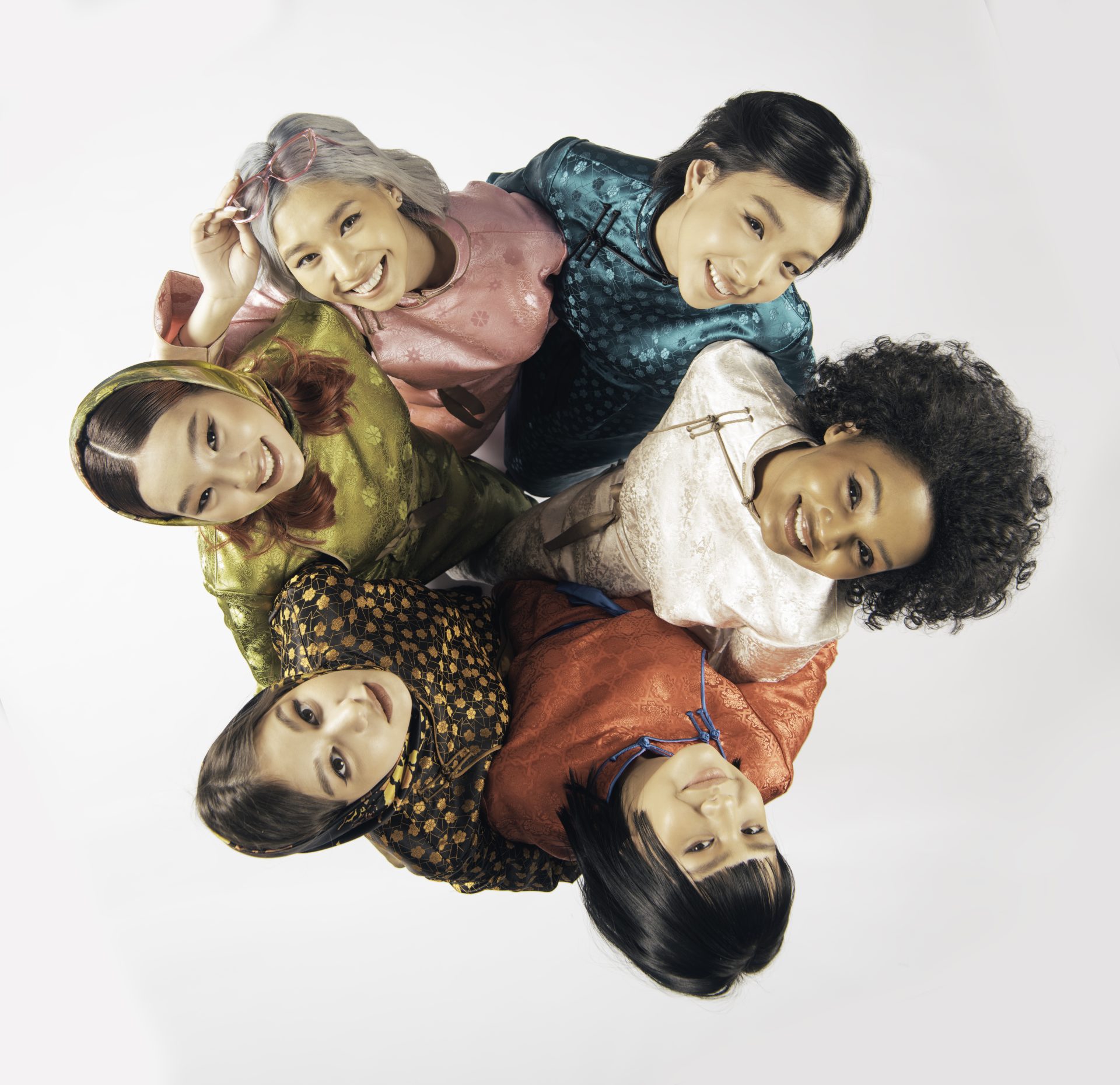 A group of six diverse women standing in a circle and looking up, wearing colorful traditional clothing. They are smiling and standing against a plain white background.