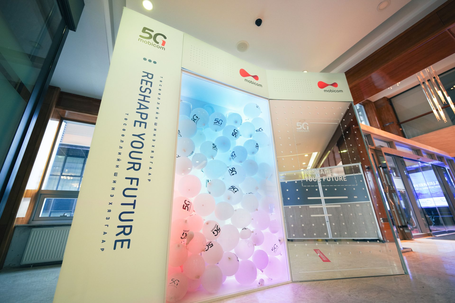 A modern exhibition booth promoting 5G technology with the slogan "Reshape Your Future." The display features a glass enclosure filled with balloons, alongside sleek, futuristic design elements and multimedia screens.