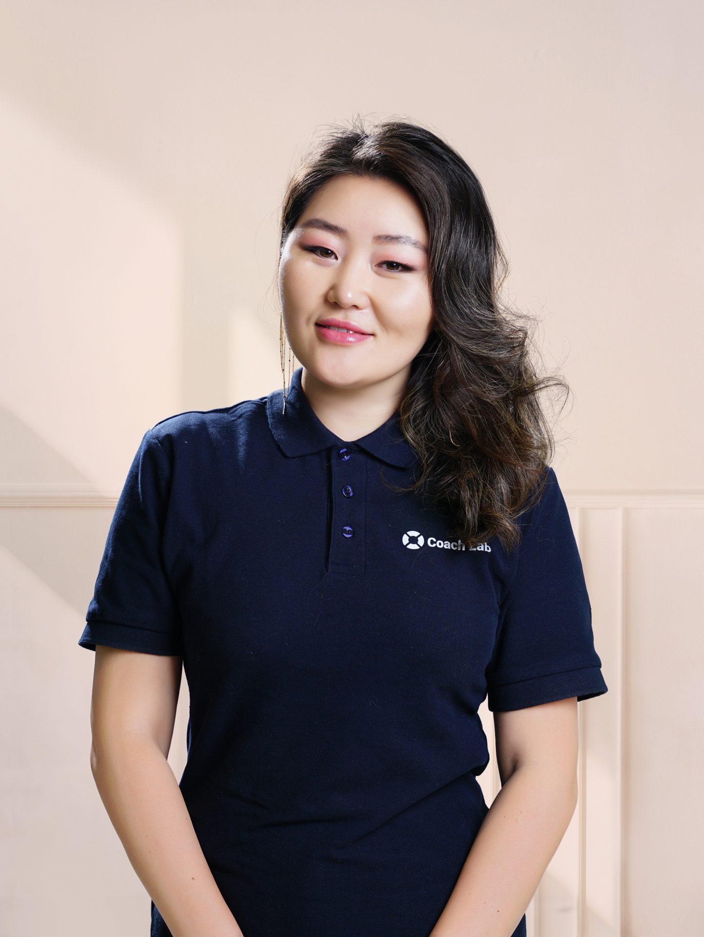 A woman with long dark hair smiles slightly. She is wearing a navy blue polo shirt with a small logo on the chest, standing against a light-colored background.