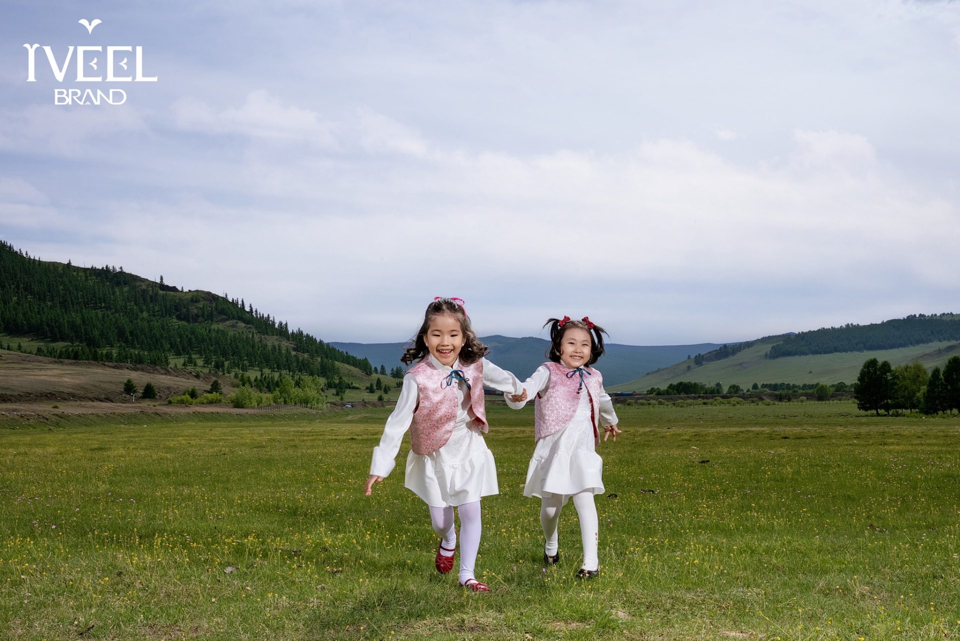 Two young girls joyfully run hand in hand across a grassy field. They wear matching pink vests and white outfits. The landscape is lush with green trees and hills under a cloudy sky. A logo is visible in the upper left corner.