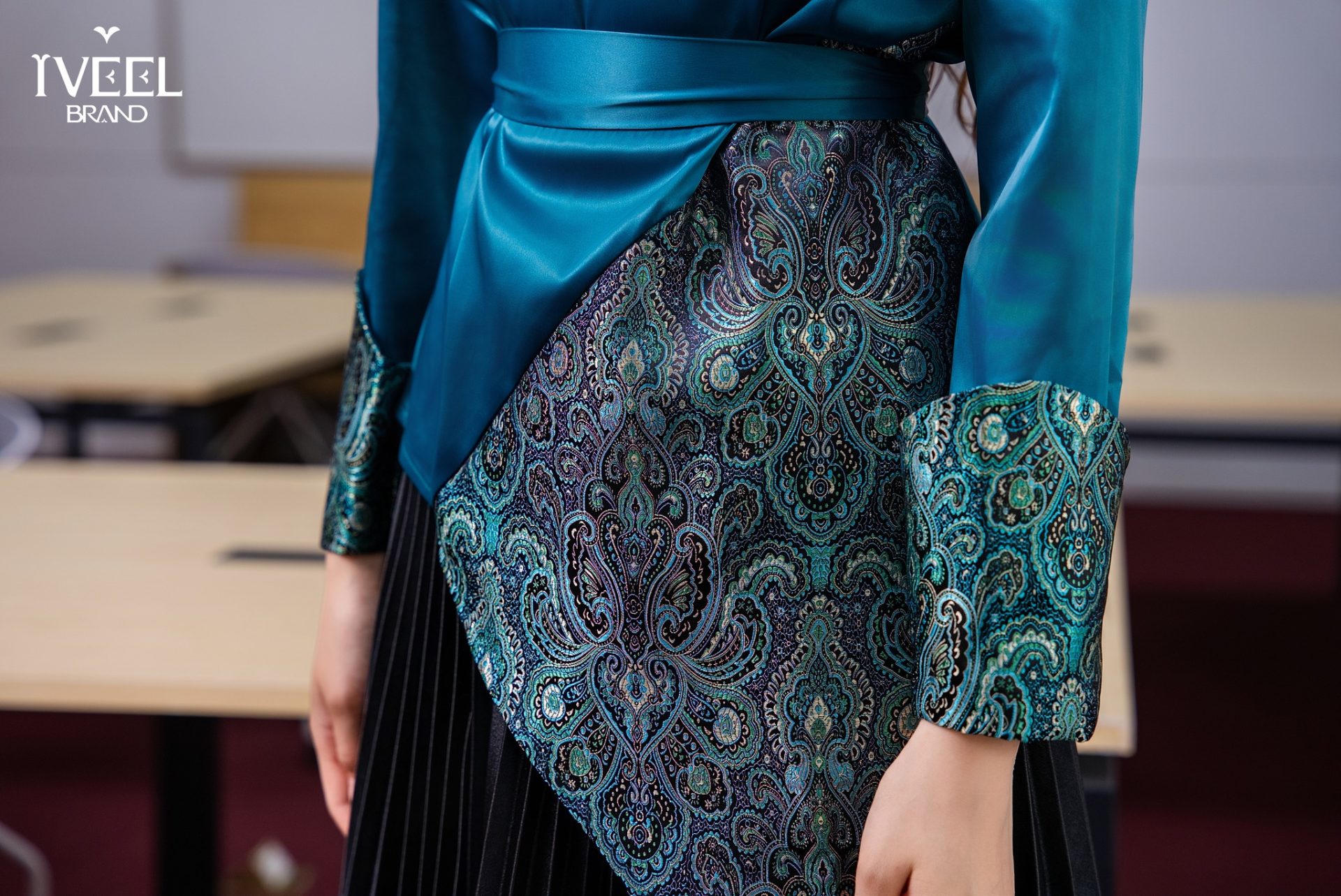 A close-up of a person wearing a teal silk blouse with an ornate paisley pattern on the lower part and sleeves. The outfit is accented with a matching silk belt, worn over a pleated black skirt. Tables are seen in the blurred background.