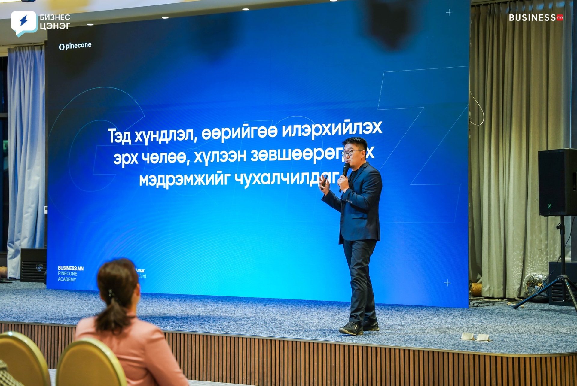 A speaker stands on a stage, speaking to an audience. A large blue screen behind them displays text in a non-Latin script. The setting appears to be a conference or business event, with attendees seated and listening.