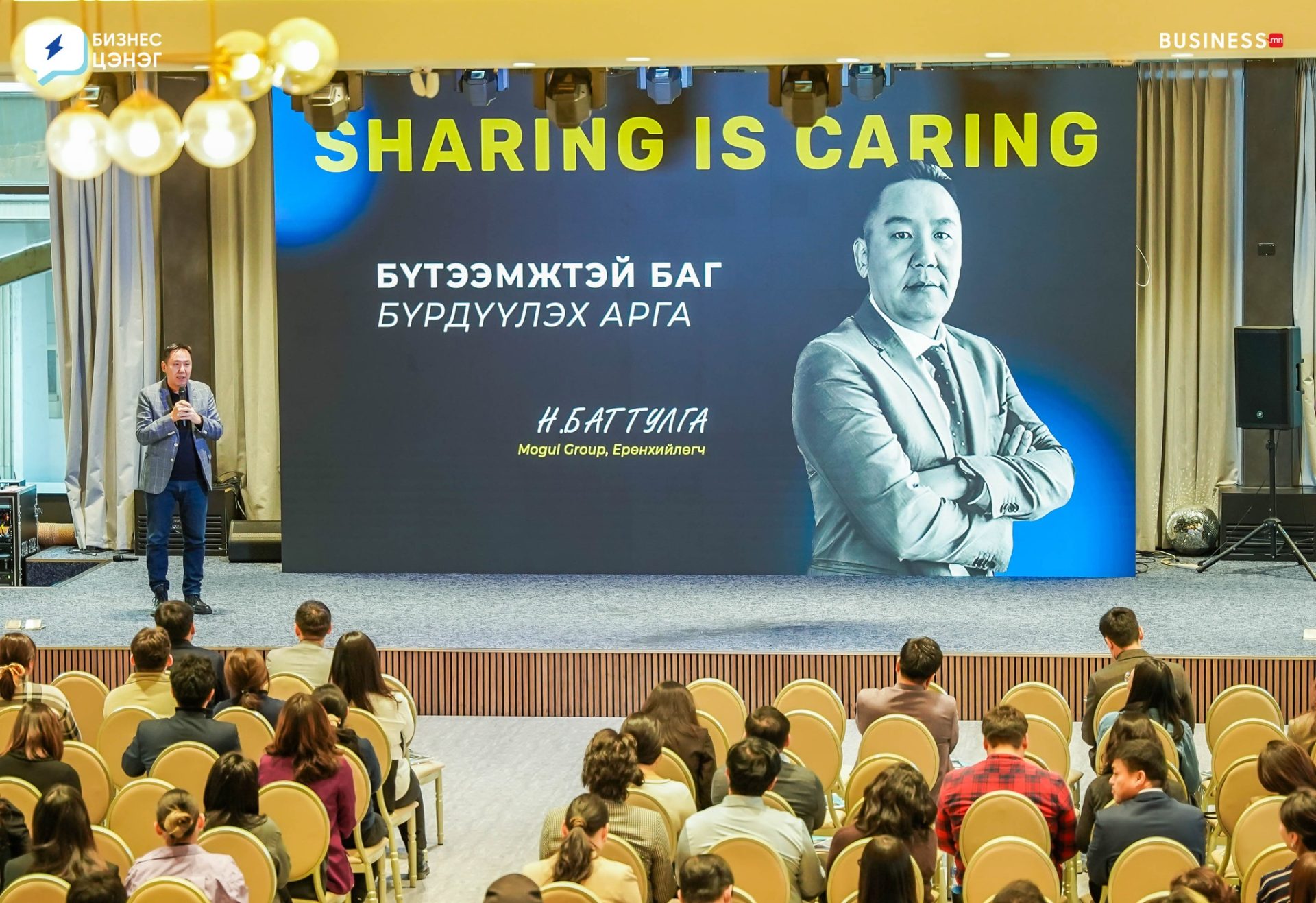 A speaker stands on stage addressing an audience in a conference room. The backdrop displays the words "Sharing is Caring" along with text in another language, beside a portrait of the speaker. Attendees are seated, listening attentively.