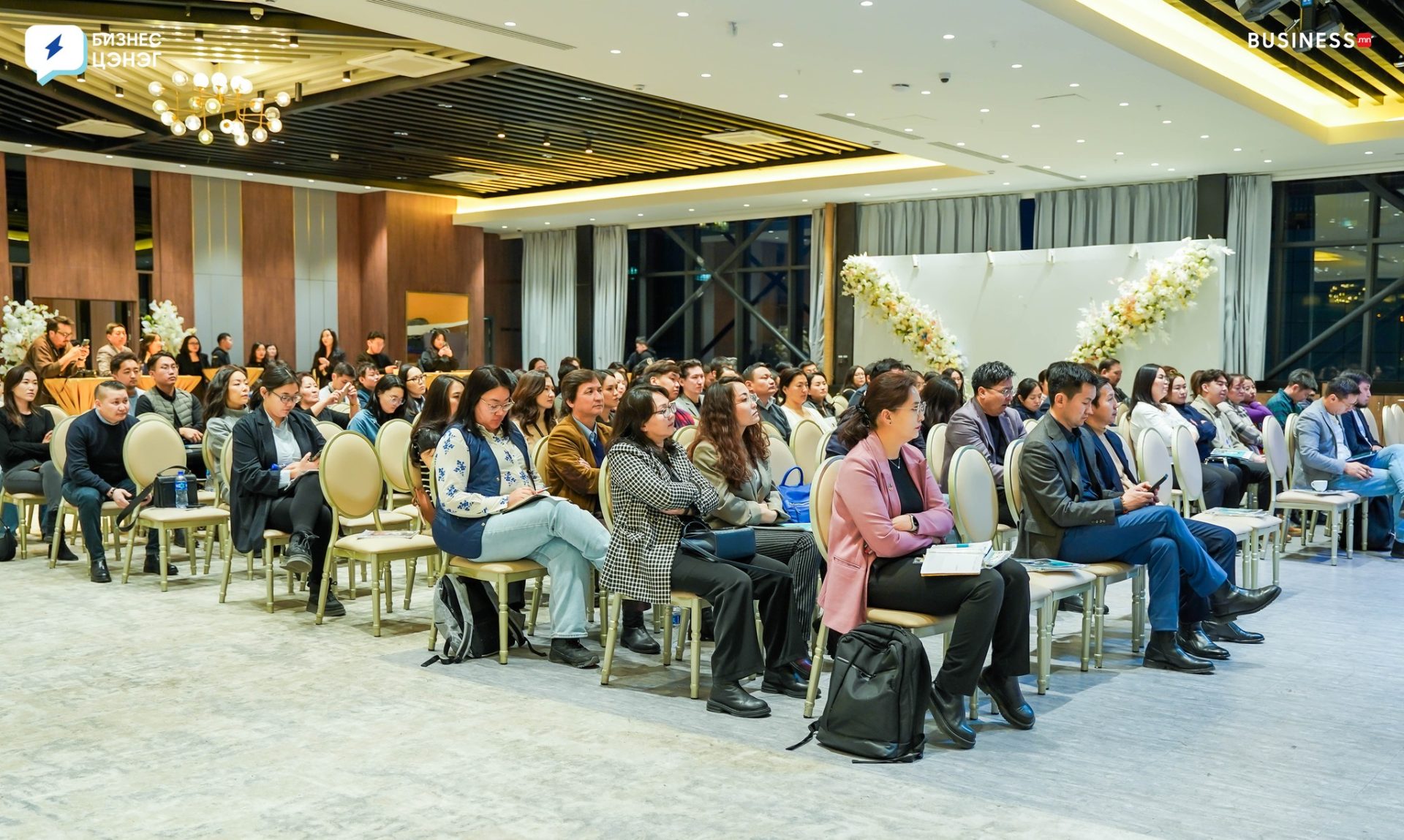 People are seated in rows in a conference room, attentively listening to a speaker. The room features modern decor with a floral arrangement at the front. Casual and business attire suggests a professional event or seminar.