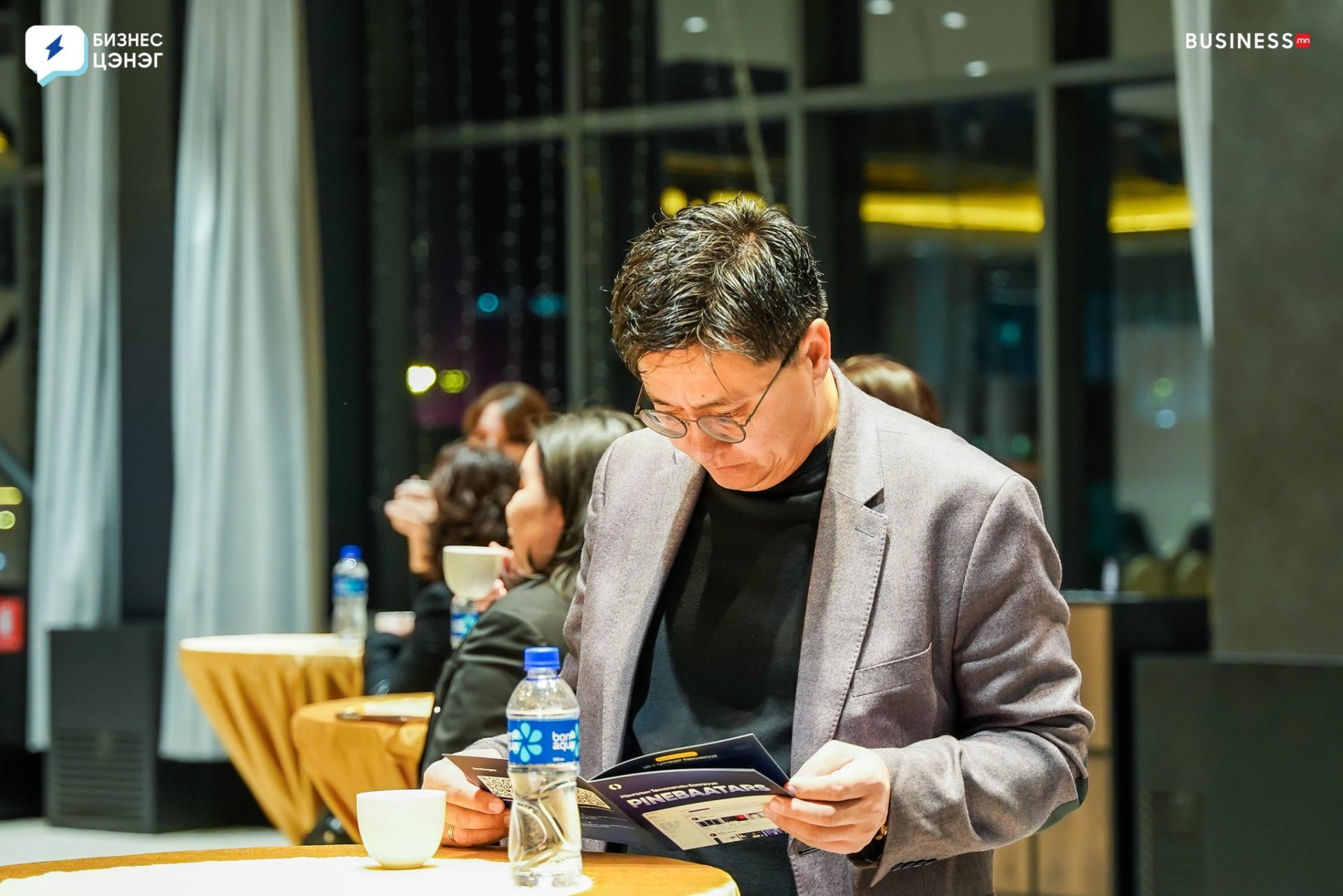 A person in a gray blazer stands at a table, reading a booklet. There's a cup and a bottle of water on the table. In the background, other people sit and engage in conversation in a brightly lit room.