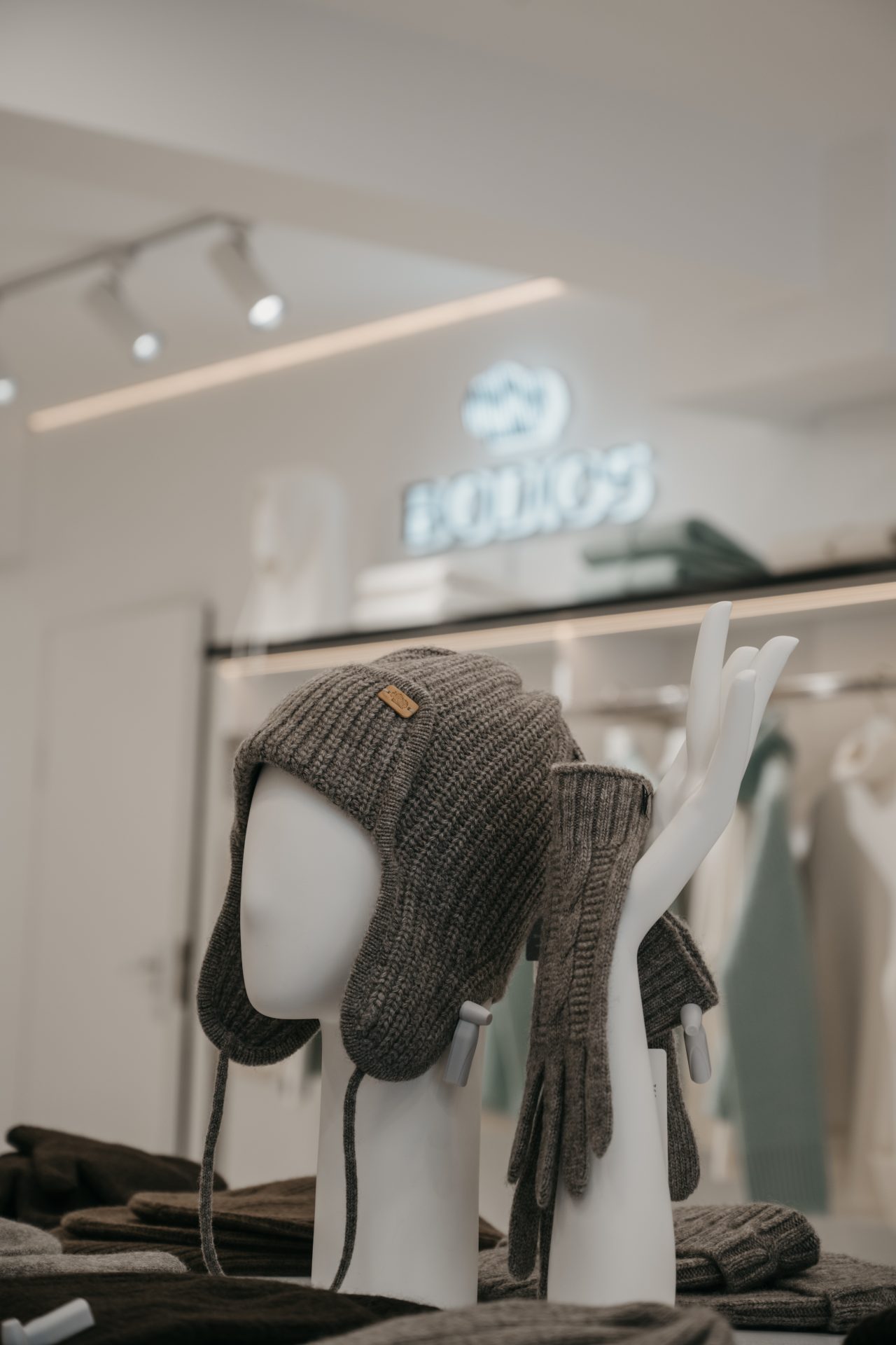 A gray knit hat with ear flaps and matching gloves are displayed on a mannequin hand in a modern store. The background shows a blurred sign and clothing racks with various garments.