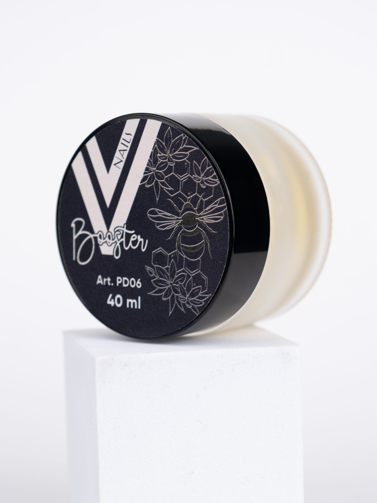 A small round jar of V Nails Booster sits on a white pedestal. The black lid features a bee and floral design, with the text "Art. PD06" and "40 ml." The jar appears slightly translucent with a light yellow hue inside.