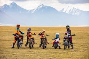 Five motocross riders, including children, in colorful gear and helmets, are on dirt bikes in a grassy field with snow-capped mountains in the background. The sky is cloudy, adding a dramatic effect to the scenic landscape.