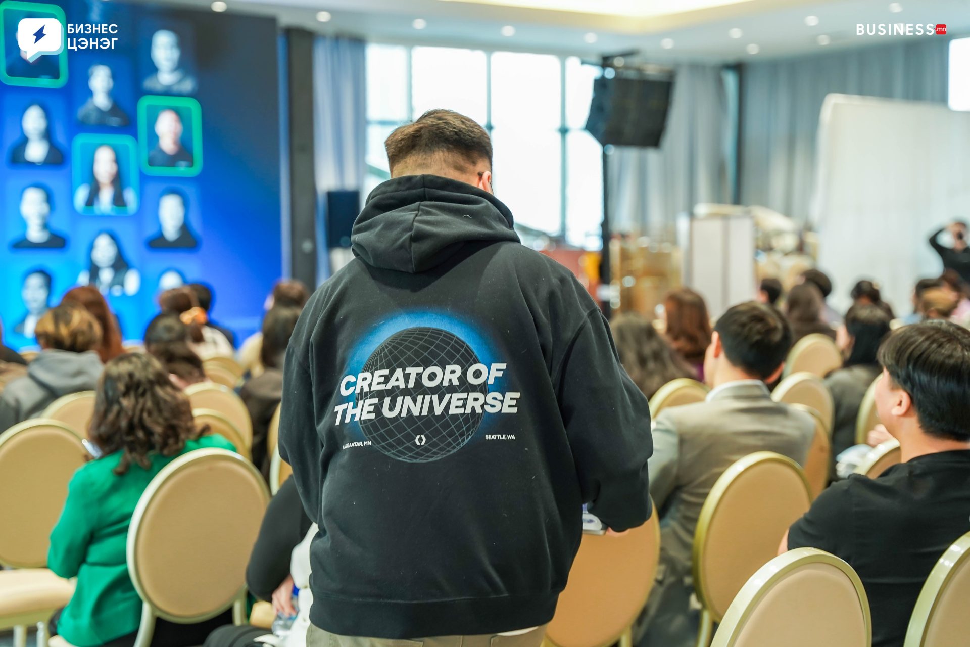 A person wearing a black hoodie with "CREATOR OF THE UNIVERSE" printed on the back stands in an auditorium. The room is filled with seated attendees facing a stage displaying a large screen with graphics and photos.