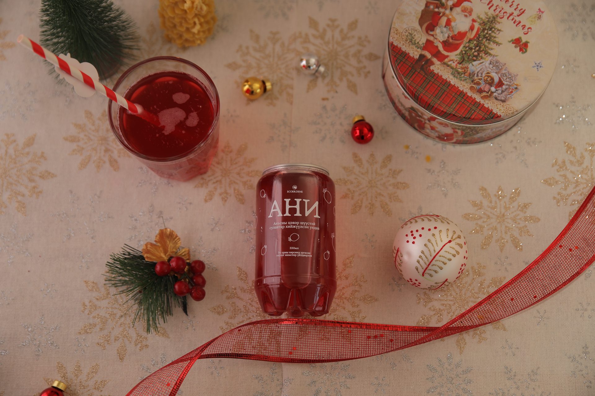 A festive arrangement featuring a red drink in a glass with a striped straw on a tablecloth with gold snowflakes. Nearby are a can with Cyrillic text, a round ornament, red ribbon, small decorations, and a holiday-themed tin.