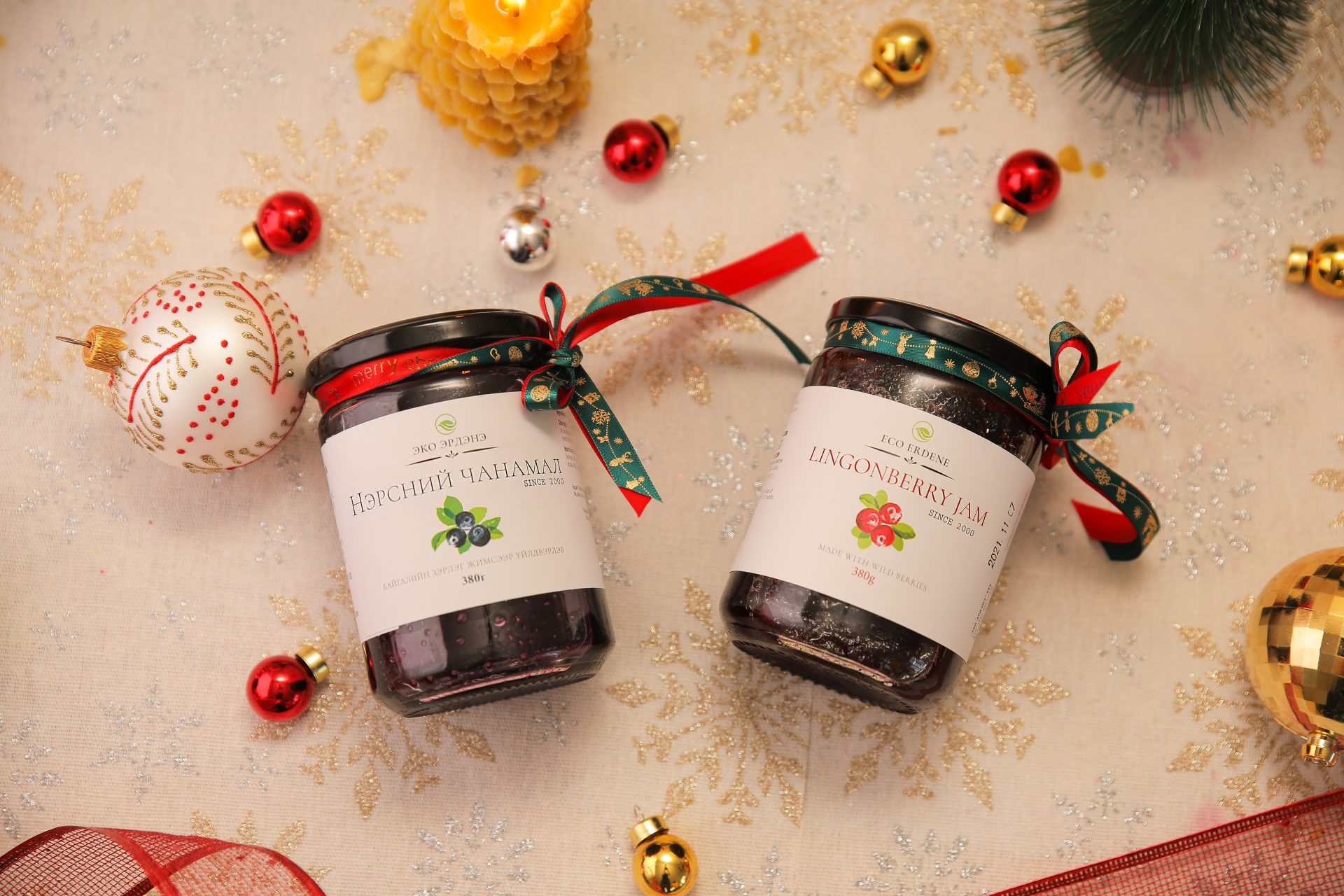 Two jars of lingonberry jam on a festive tablecloth with snowflake designs, surrounded by holiday decorations: red and gold ornaments, a beaded white ornament, a golden candle, and a green pine decoration.