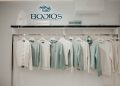 A clothing rack displays a collection of white and light green sweaters and cardigans in a store. The wall above showcases the brand name "BODIOS" in illuminated letters. The atmosphere is bright and minimalist.