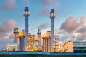 A large industrial power plant with multiple tall chimneys releases light smoke against a backdrop of a cloudy sky at dusk. Illuminated by bright lights, the facility stands out in the эрчим хүчний салбарт гэрэл асаах мэдээ, showcasing its structures and machinery.