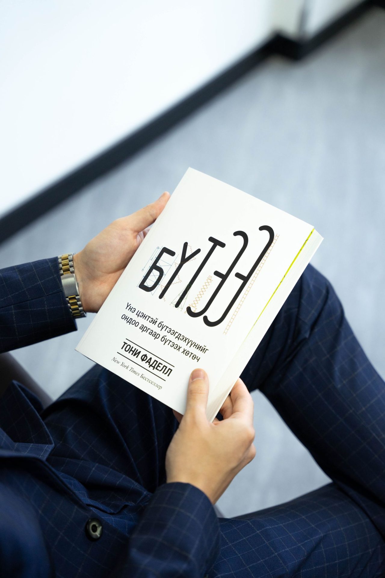 A person in a blue suit holding an open book with a white cover and text in what appears to be Cyrillic script. The book's cover features modern design elements and typography. The background is a plain, light-colored wall.